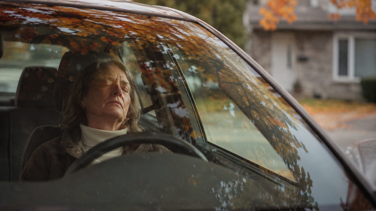 Une femme épuisée, les yeux fermés, assise dans sa voiture stationnée devant une maison, des feuilles d'automne se reflétant dans le pare-brise.