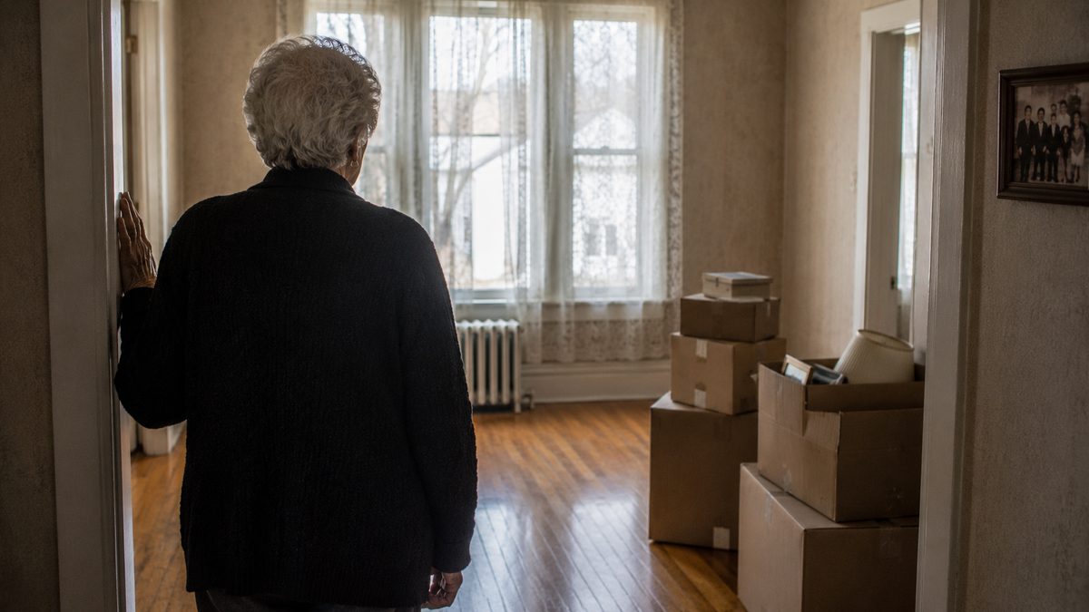Une dame âgée vue de dos dans le salon presque vide de sa maison, des boîtes de déménagement empilées près de la fenêtre.
