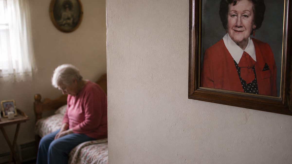 Une dame âgée assise seule sur son lit, la tête baissée, près d'un portrait encadré accroché au mur.