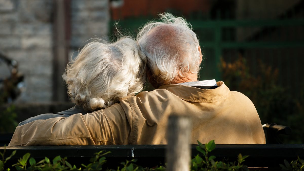 Un couple d'aînés vu de dos sur un banc de parc, la tête de la dame appuyée sur l'épaule de son compagnon.